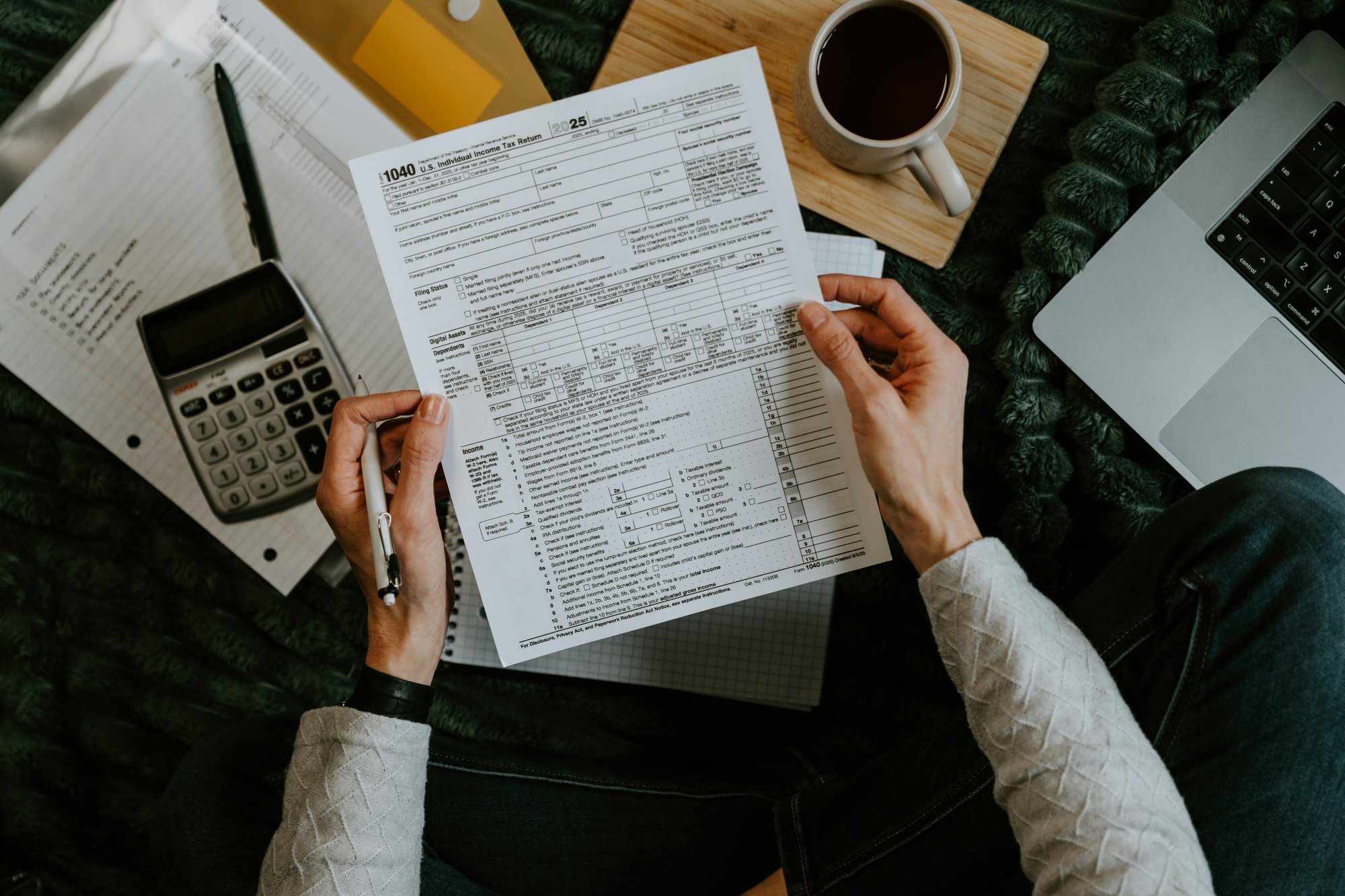 Person reviewing tax documents with a calculator, laptop, and coffee during tax preparation.